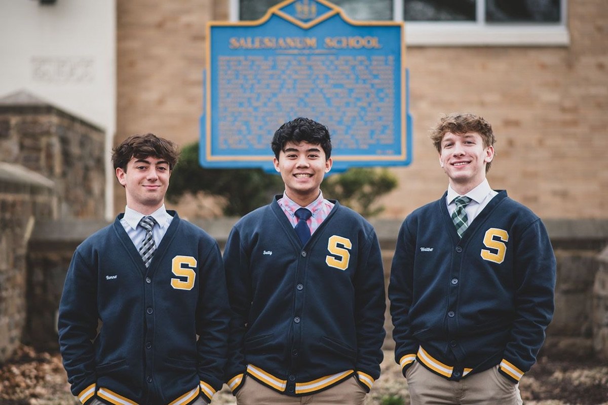 Three Salesianum students in navy letter jackets with the gold S, in front of the school’s historical marker