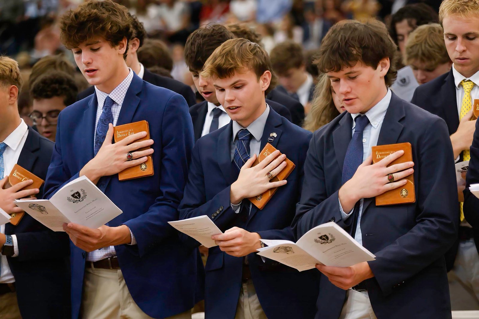 Students in navy blazers holding programs with hands over their hearts during a Mass